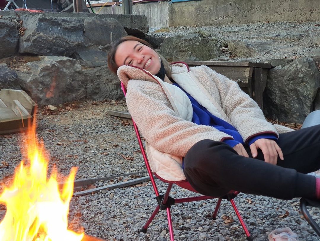야외 캠핑장에서 불 앞에 눕듯이 앉아 해사하게 웃고 있다.